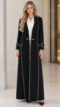 Front full-length view of woman wearing black tailored maxi duster coat with white contrast piping and gold buttons